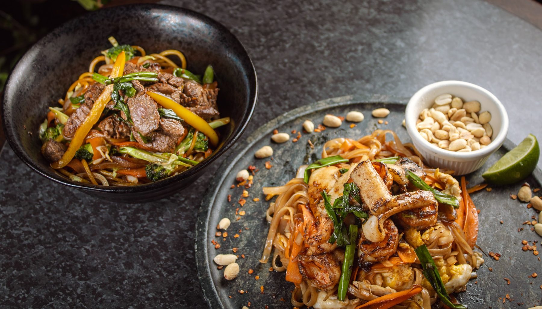 Yakisoba com carne na tigela e pad thai com camarão no prato de pedra, com amendoim e limão