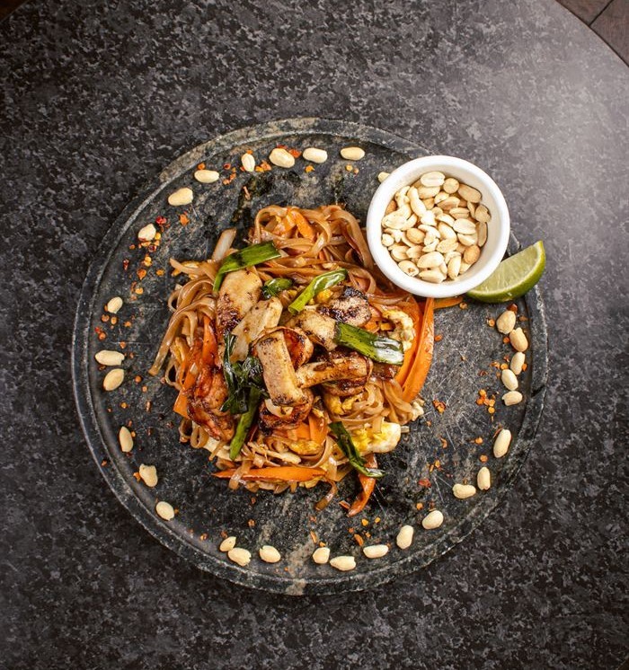 Pad thai servido em prato de pedra com amendoim, limão e castanhas